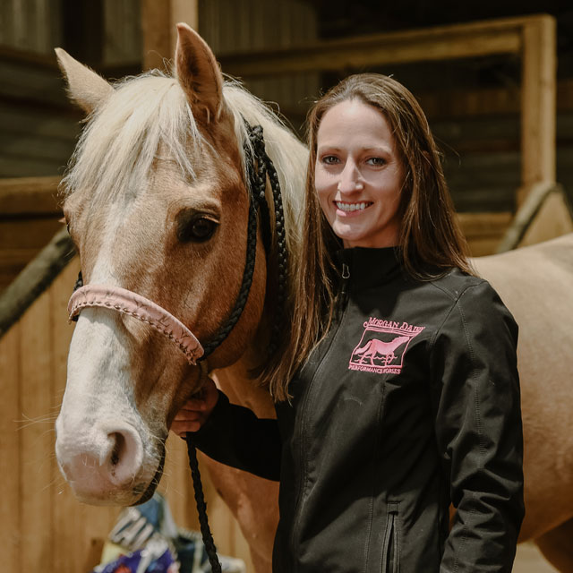 Amanda Morgan de Morgan Dairy Performance Horses vistiendo una chaqueta negra y de pie junto a su caballo rojo.